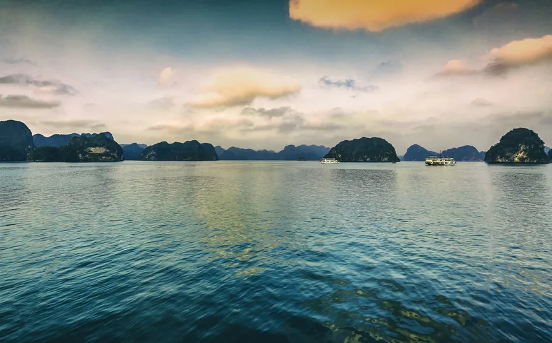 Những du thuyền lướt nhẹ giữa các đảo đá vôi trên mặt nước yên bình của vịnh Hạ Long lúc hoàng hôn.