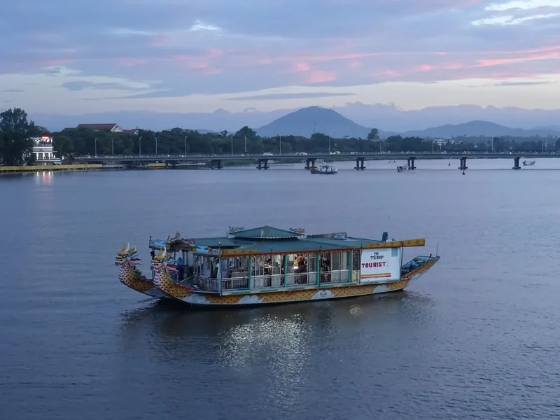 Thuyền rồng du lịch trên sông Hương ở Huế lúc hoàng hôn, phía sau là cầu và núi.