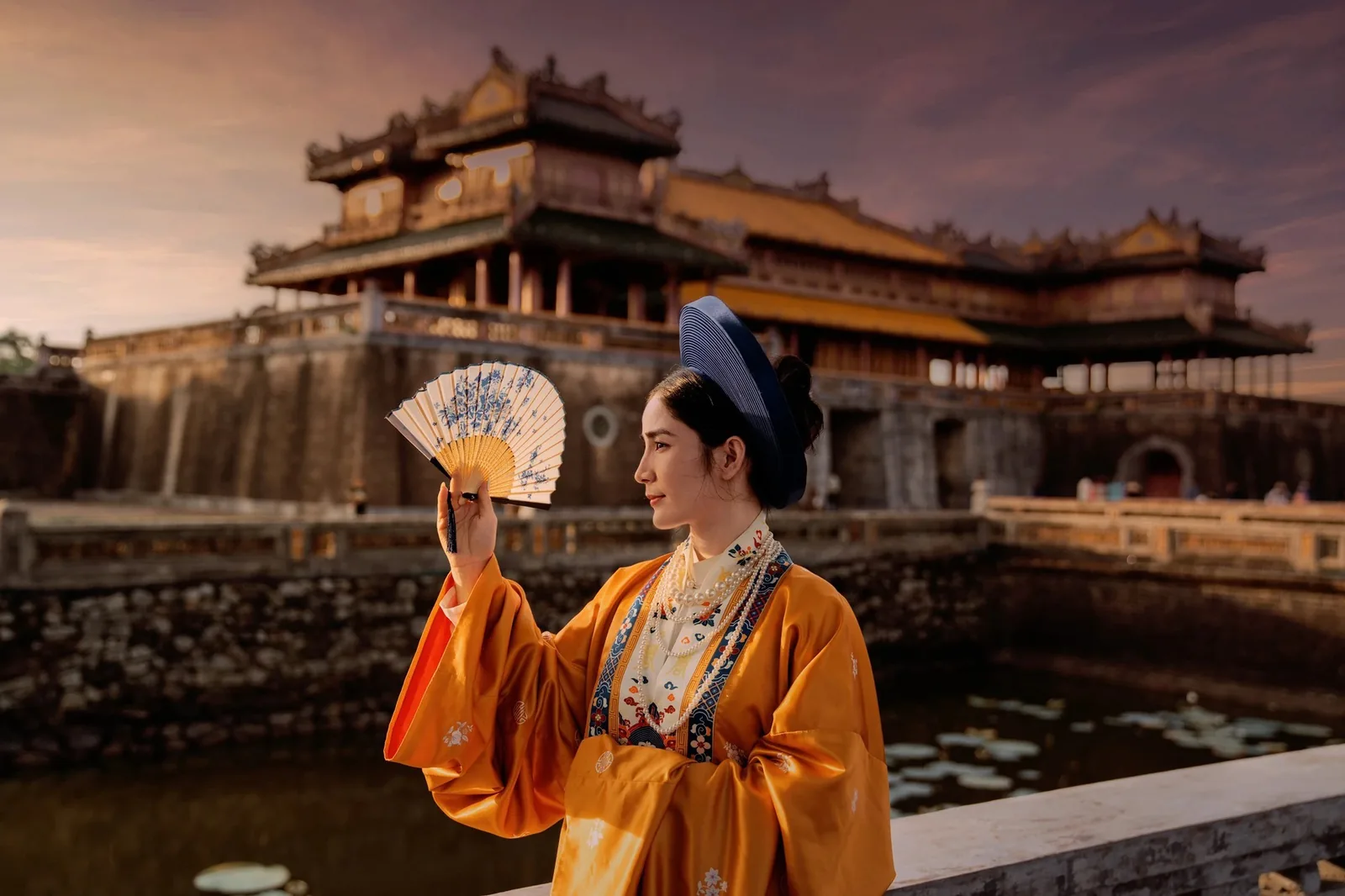 Người phụ nữ trong áo dài vàng cầm quạt đứng trước cổng Ngọ Môn của Đại Nội Huế lúc hoàng hôn.